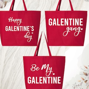 May include: Three red tote bags with white text. The bags read "Happy Galentine's Day", "Galentine gang", and "Be My Galentine". Each bag has long handles and a rectangular shape. The bags are on a white textured surface.