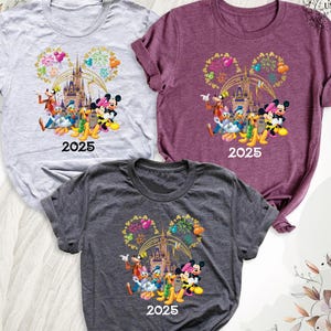 Puede incluir: Tres camisetas con un estampado gr&aacute;fico de Mickey Mouse y sus amigos frente al Castillo de Cenicienta con fuegos artificiales. El texto "2025" est&aacute; impreso en cada camiseta. Las camisetas son de color gris, morado y gris oscuro.