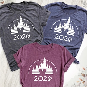 May include: Three short-sleeved t-shirts in charcoal, navy, and burgundy. Each shirt features a white graphic of a castle with Mickey Mouse ears and the year "2026" printed below. The shirts are made of a soft material.