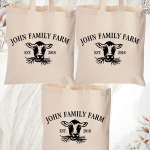 May include: Three white canvas tote bags with black handles and a black graphic of a cow's head with the text "JOHN FAMILY FARM EST. 2018" printed on each bag.