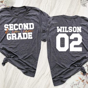 May include: Two dark gray t-shirts. The front shirt says "SECOND GRADE" with "dream team" in colorful script. The back shirt says "WILSON 02" in white block letters. The shirts are knotted at the bottom.