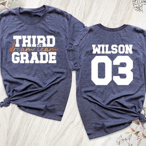May include: Two navy blue t-shirts. The front of one shirt reads "THIRD dream team GRADE" in white and orange lettering. The back of the other shirt says "WILSON 03" in white. Both shirts are knotted at the bottom.