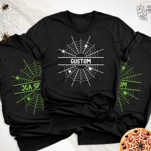 May include: A black short-sleeved t-shirt featuring a white spiderweb graphic centered on the chest. A horizontal white bar across the middle of the web contains the text CUSTOM in a bold, sans-serif font. Small white spiders are visible on the web.