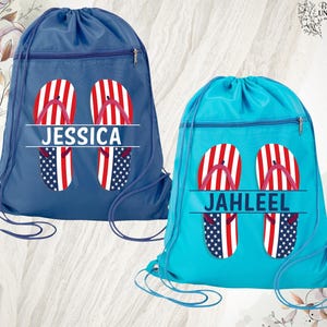 May include: Two drawstring backpacks, one navy blue and one turquoise. Each bag features a design of red and white striped flip-flops with a blue and white star pattern. The navy bag says "JESSICA" and the turquoise bag says "JAHLEEL".