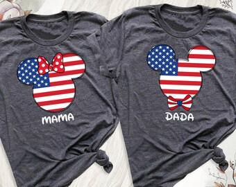 May include: Two dark gray t-shirts with a patriotic Mickey Mouse design. One shirt says "MAMA" and features a red bow. The other says "DADA" and has a bow tie. The Mickey Mouse design incorporates the American flag.