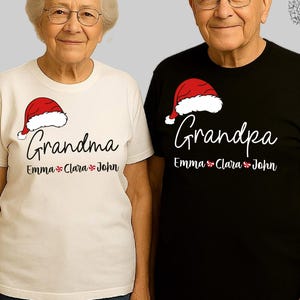以下が含まれることがあります： お揃いのクリスマスTシャツを着た2人。片方のシャツは白で、「Grandma」の文字とサンタ帽のグラフィックが入っています。もう一方のシャツは黒で、「Grandpa」の文字とサンタ帽のグラフィックが入っています。どちらのシャツにも「Emma, Clara, John」という名前が書かれています。