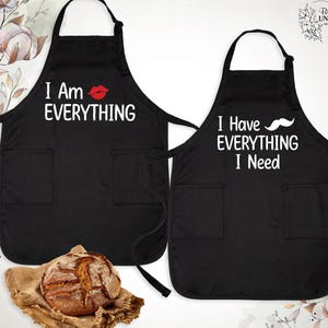 May include: Two black aprons with white text. One apron says "I Am Everything" with a red lip print. The other apron says "I Have Everything I Need" with a black mustache print.