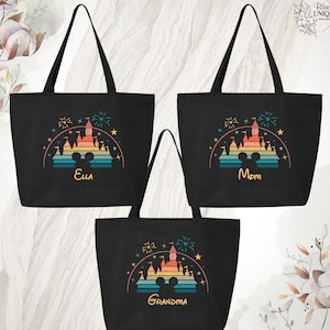 May include: Three black canvas tote bags with a retro rainbow design of a castle with fireworks and a Mickey Mouse silhouette. The bags are personalized with the names "Ella", "Mom", and "Grandma".