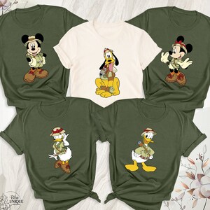 May include: Four green and white t-shirts with different Disney characters wearing safari hats and outfits. The characters are Mickey Mouse, Pluto, Donald Duck, and Daisy Duck.