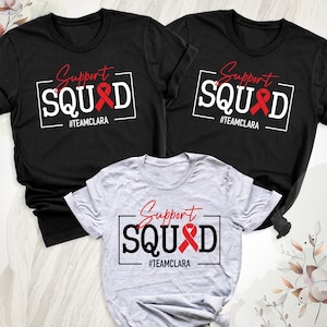 May include: Three t-shirts, two black and one gray, are displayed. Each shirt features the words "Support SQUAD" in a white box with a red ribbon and the hashtag #TEAMCLARA. The text is in a handwritten style font.