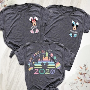 May include: Three dark gray t-shirts with Easter-themed designs. One shirt features Minnie Mouse with bunny ears and the name "Olivia." Another has Mickey Mouse with bunny ears and the name "Christian." The third shirt has "Happy Easter 2020" with a castle and rainbow.