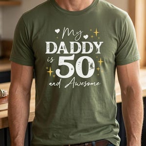 May include: An olive green short-sleeved t-shirt featuring white and yellow graphic text that reads My DADDY is 50 and Awesome, decorated with small white hearts and yellow four-pointed stars arranged around the central text.