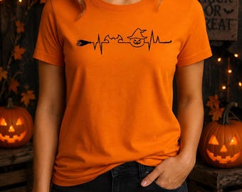 May include: A bright orange short-sleeved t-shirt featuring a black graphic print of a heartbeat line. The design incorporates a broomstick, a bat, and a jack-o'-lantern wearing a witch hat integrated into the EKG pulse pattern.
