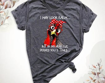Peut inclure: Un t-shirt gris chiné foncé à manches courtes avec un motif de poulet portant un foulard rouge à pois blancs. Le texte indique : I MAY LOOK CALM... BUT IN MY HEAD I'VE PECKED YOU 3 TIMES... Le t-shirt présente une encolure ronde.