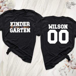 以下が含まれることがあります： 黒いTシャツ2枚。1枚には、白、オレンジ、青、紫で「KINDER dream team GARTEN」と書かれています。もう1枚には、白で「WILSON 00」と書かれています。どちらのシャツも裾で結ばれており、カジュアルな印象です。