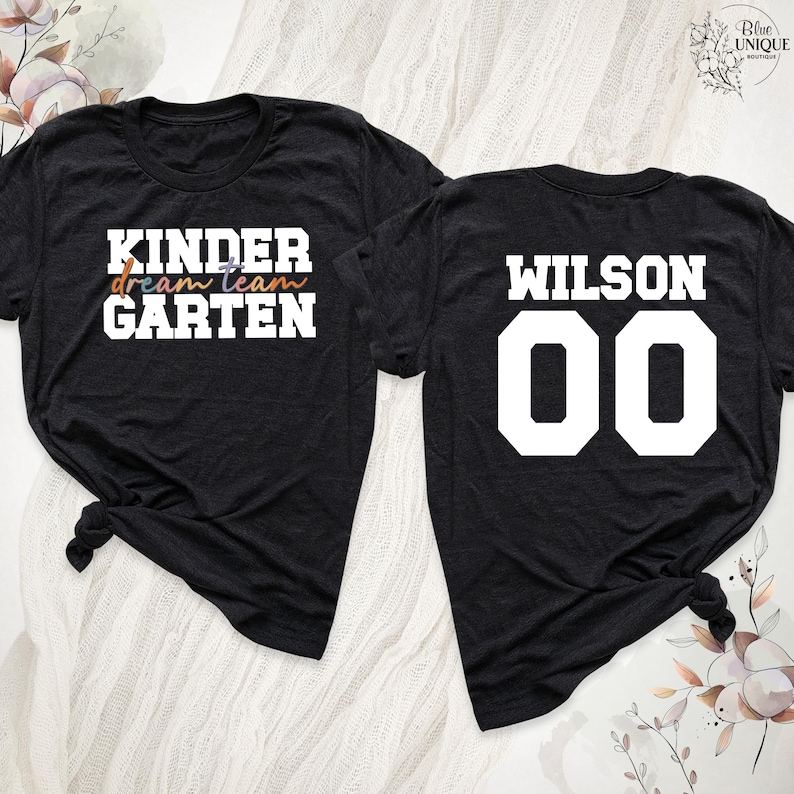 以下が含まれることがあります： 黒いTシャツ2枚。1枚には、白、オレンジ、青、紫で「KINDER dream team GARTEN」と書かれています。もう1枚には、白で「WILSON 00」と書かれています。どちらのシャツも裾で結ばれており、カジュアルな印象です。