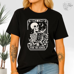 May include: A black t-shirt featuring a white graphic of a skeleton reading a book within a rectangular frame. The design includes moon phases, stars, and leaves, with the text THE READER printed at the bottom of the graphic illustration.