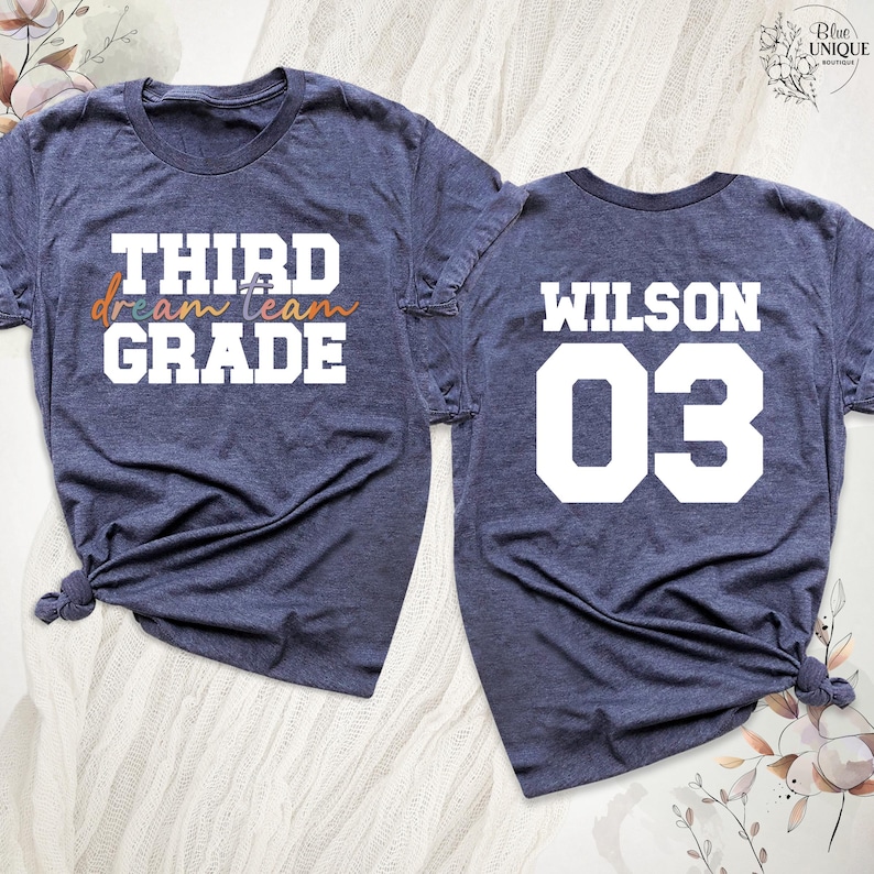 以下が含まれることがあります： 紺色のTシャツ2枚。1枚には白とオレンジ色の文字で「THIRD dream team GRADE」と書かれています。もう1枚には白で「WILSON 03」と書かれています。どちらも裾が結ばれています。学校やスポーツに最適です。