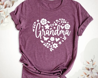 May include: A heathered maroon short-sleeve t-shirt featuring a large white graphic of a heart shape composed of various flowers, leaves, stars, and a butterfly. The word Grandma is printed in a white script font across the center of the heart.