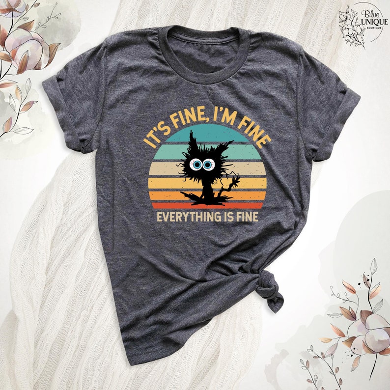 May include: A dark grey t-shirt with a vintage-style sunset graphic featuring a black cat with large blue eyes. The text "It's fine, I'm fine, everything is fine" is printed on the shirt.