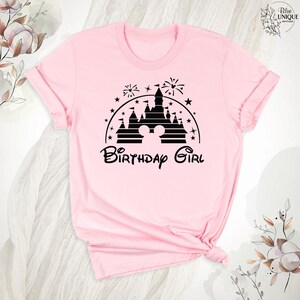 Pode incluir: Camiseta rosa com uma silhueta preta do castelo da Cinderela e orelhas do Mickey Mouse. O texto "Birthday Girl" est&aacute; abaixo do castelo.
