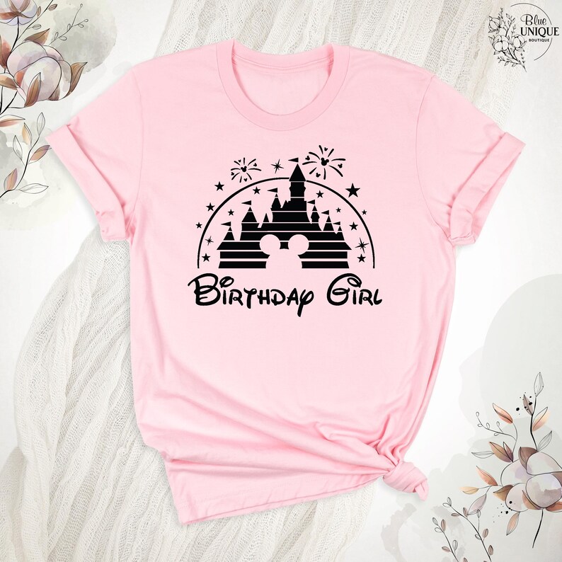 Pode incluir: Camiseta rosa com uma silhueta preta do castelo da Cinderela e orelhas do Mickey Mouse. O texto "Birthday Girl" est&aacute; abaixo do castelo.