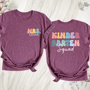 May include: Two maroon t-shirts with rolled sleeves. One shirt has "MRS. Olivia" in colorful letters. The other shirt has "KINDERGARTEN Squad" in colorful letters. Both shirts are knotted at the bottom.