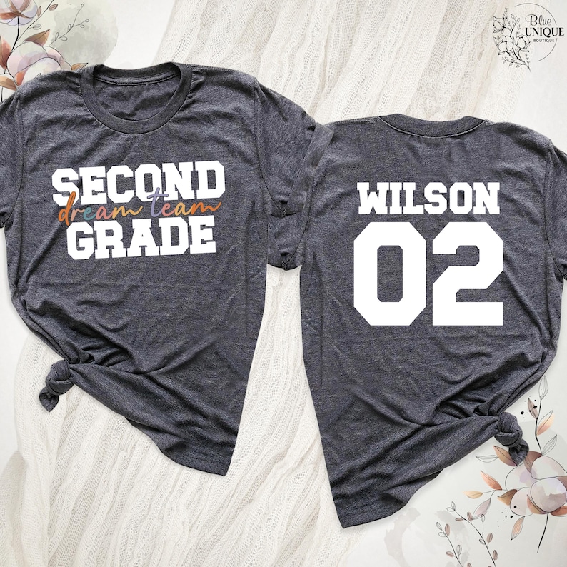 以下が含まれることがあります： 2枚の濃いグレーのTシャツ。前面のシャツには、カラフルなスクリプトで「dream team」と「SECOND GRADE」と書かれています。背面のシャツには、白いブロック文字で「WILSON 02」と書かれています。シャツは下部で結ばれています。