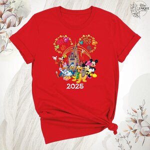 Puede incluir: Camiseta roja con un gr&aacute;fico de Mickey Mouse, Minnie Mouse, Donald Duck, Goofy y Pluto frente al Castillo de Cenicienta con fuegos artificiales. El texto "2025" est&aacute; debajo de los personajes.