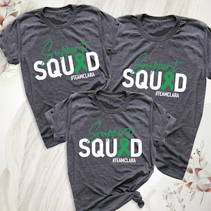 May include: Three charcoal gray short-sleeve t-shirts featuring the text Support SQUAD in white and green, with a green awareness ribbon replacing the letter A. The text #TEAMCLARA is printed in white below the main graphic on each shirt.