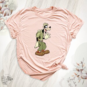 May include: A pink t-shirt with a cartoon image of Goofy dressed as a park ranger. Goofy is wearing a green shirt, brown pants, a brown hat, and a brown backpack. He is waving with his right hand.