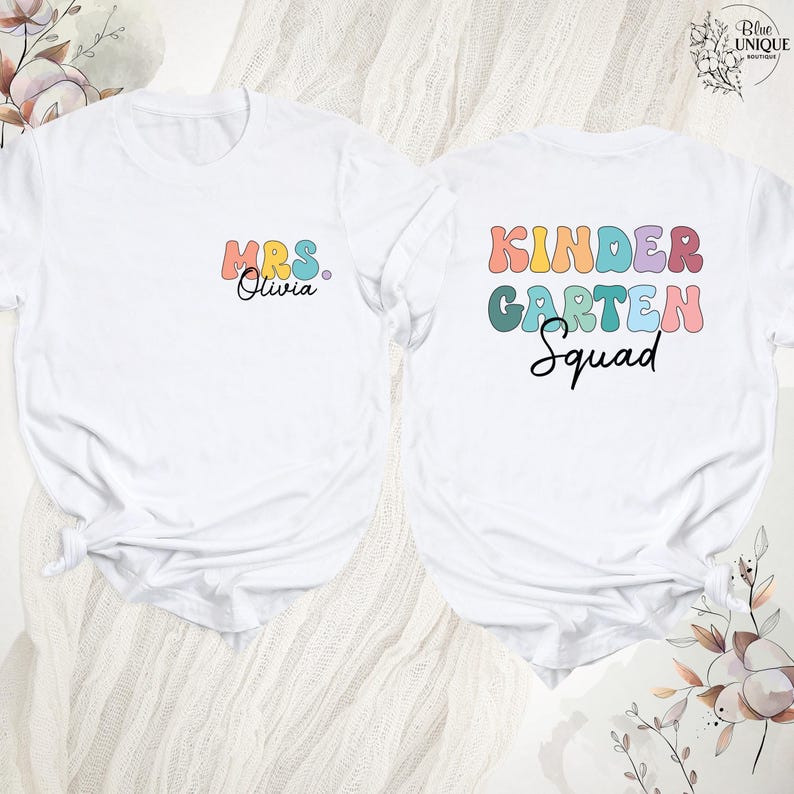 May include: Two white t-shirts with colorful text. One shirt says "Mrs. Olivia" in retro font. The other says "Kindergarten Squad" in a similar style. Both shirts are likely for a teacher or school staff.