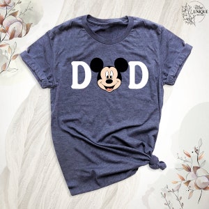 May include: Navy blue t-shirt with the word "DAD" and a Mickey Mouse graphic. The shirt has a crew neck and short sleeves. The design is printed in white and black.