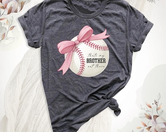 May include: A dark heather gray short-sleeve t-shirt featuring a graphic of a white baseball adorned with a large pink ribbon bow. The text on the baseball reads, "that's my BROTHER out there" in black lettering. The shirt has a crew neckline.