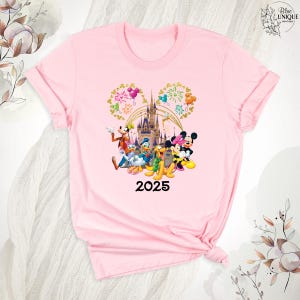Puede incluir: Camiseta rosa con un gr&aacute;fico de Mickey Mouse y sus amigos frente al Castillo de Cenicienta con fuegos artificiales. El texto "2025" est&aacute; debajo del gr&aacute;fico.