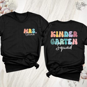 May include: Two black t-shirts with colorful text. One shirt says "MRS. Olivia" in pastel colors. The other says "KINDERGARTEN Squad" in a retro font. The shirts are knotted at the bottom.