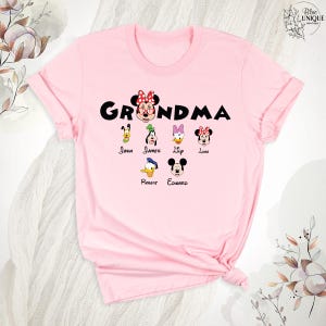 Puede incluir: Camiseta rosa con el texto "Grandma" en letras negras. La camiseta presenta una imagen de dibujos animados de Minnie Mouse con un lazo y otros seis personajes de Disney, cada uno con un nombre debajo: John, James, Lily, Luna, Robert, Edward.
