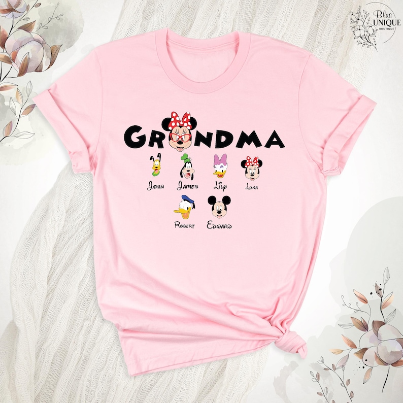 Puede incluir: Camiseta rosa con el texto "Grandma" en letras negras. La camiseta presenta una imagen de dibujos animados de Minnie Mouse con un lazo y otros seis personajes de Disney, cada uno con un nombre debajo: John, James, Lily, Luna, Robert, Edward.