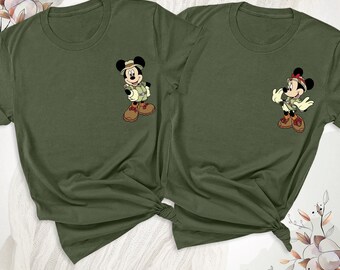 May include: Two olive green crew neck t-shirts. The left shirt features a graphic of Mickey Mouse dressed in safari gear with a hat and boots. The right shirt features a graphic of Minnie Mouse in matching safari attire with a red bow.