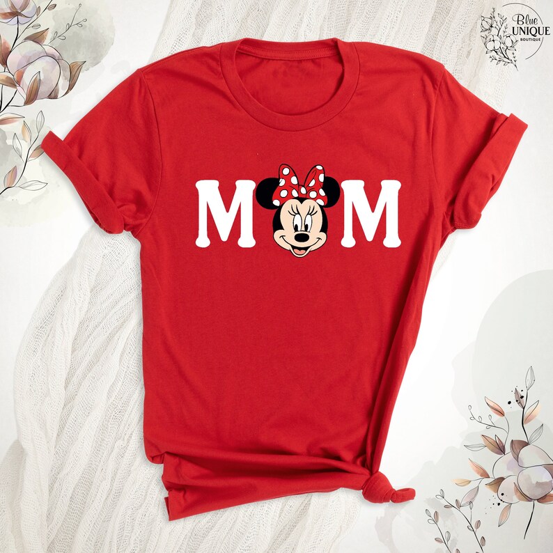May include: A red t-shirt with the word "MOM" in white, with Minnie Mouse's face as the "O". Minnie Mouse has a red bow with white spots. The shirt is folded. The image has a floral design in the top right corner.