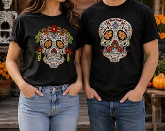 May include: Two black t-shirts featuring colorful sugar skull graphics. The left shirt displays a skull adorned with flowers and dangling earrings. The right shirt features a skull with a mustache and intricate decorative patterns.