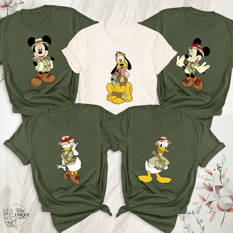 May include: Set of four green t-shirts with cartoon images of Mickey Mouse, Pluto, Donald Duck, and Daisy Duck dressed in safari outfits. Each character is wearing a safari hat and carrying a backpack.