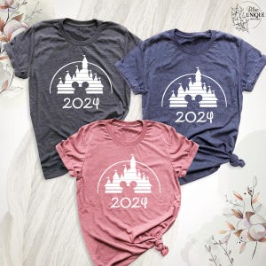 May include: Three short-sleeved t-shirts in charcoal, navy blue, and rose colours. Each shirt features a white graphic of a castle with Mickey Mouse ears and the number 2024. The shirts are displayed on a white, textured surface.