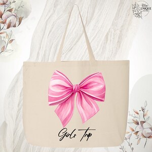 May include: A beige tote bag featuring a large pink bow illustration with white accents. The words "Girls Trip" are written in a script font below the bow. The bag has two handles and is made of a canvas-like material.