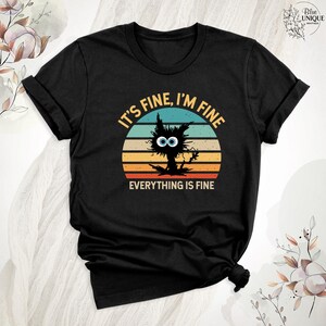 May include: Black t-shirt with a vintage-style graphic of a cat with big eyes and the text "It's fine, I'm fine, everything is fine".