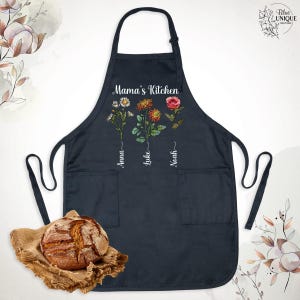 May include: Navy blue apron with the text "Mama's Kitchen" and three floral designs. The names "Anna", "Luke", and "Noah" are written below the flowers. The apron has two pockets and a bread loaf on a burlap cloth.