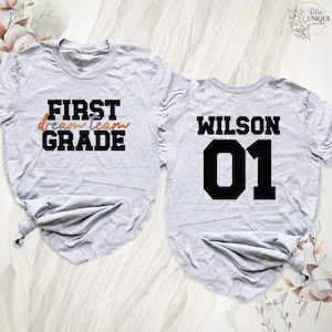 以下が含まれることがあります： 2枚のライトグレーのTシャツ。1枚のシャツには、黒いブロック体の文字で「FIRST GRADE」と、カラフルなスクリプトで「dream team」という文字が書かれています。もう1枚のシャツには、黒いブロック体の文字で「WILSON」と数字の「01」が書かれています。
