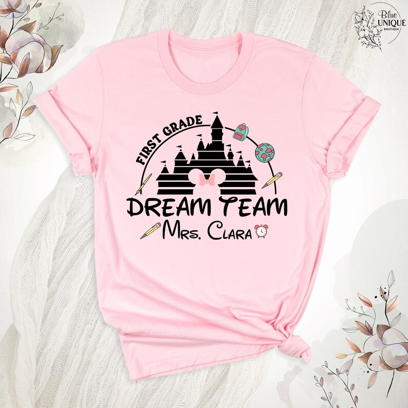 May include: A light pink t-shirt featuring a black graphic design for a first-grade teacher. The design includes a castle, pencils, a backpack, a globe, and the text "DREAM TEAM Mrs. Clara".