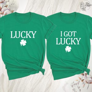 K&ouml;nnte beinhalten: Zwei gr&uuml;ne T-Shirts mit wei&szlig;em Text und einem vierbl&auml;ttrigen Kleeblatt. Ein Shirt sagt "LUCKY" und das andere "I GOT LUCKY". Die Shirts sind aus weichem Material, ideal f&uuml;r St. Patrick's Day.