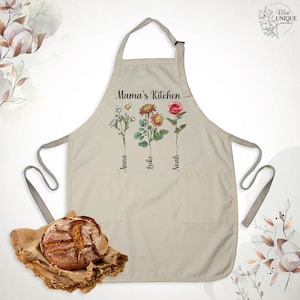 May include: Beige apron with the text "Mama's Kitchen" and floral illustrations. The apron features three flowers with the names "Anna", "Luke", and "Noah" below them. It has two pockets and a neck strap. A loaf of bread rests on a burlap cloth.